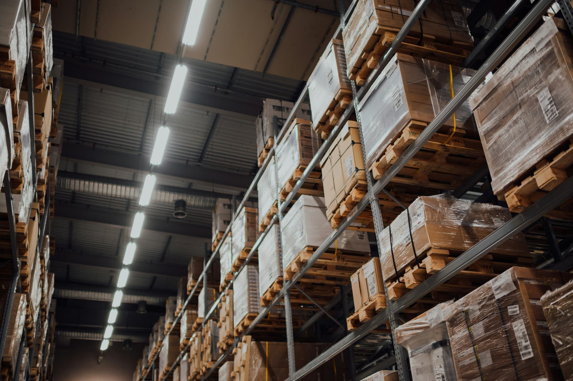Tall warehouse racking stacked with wrapped pallets of boxed goods, photographed from below under strip lighting.