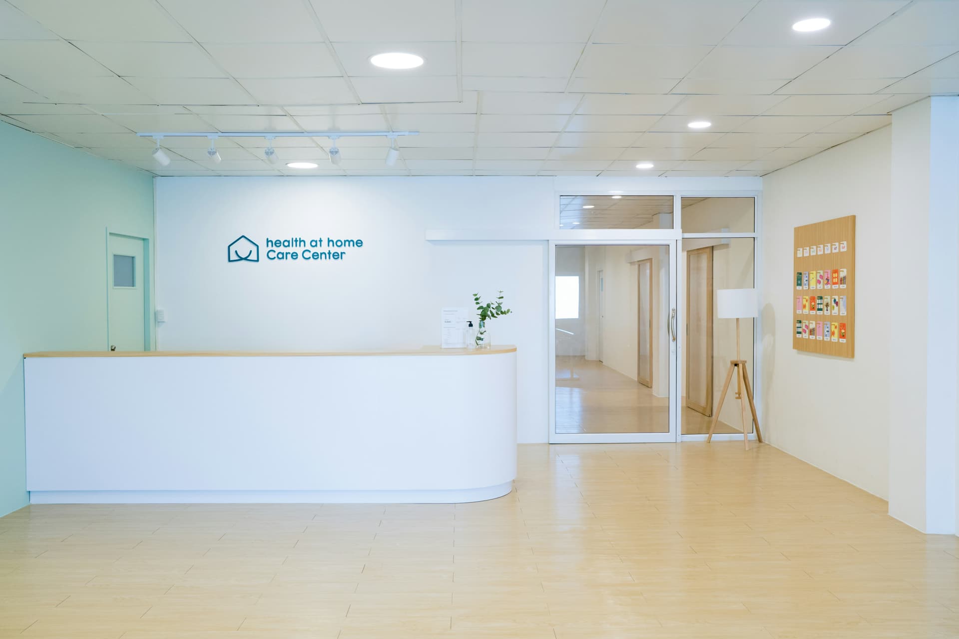 An empty reception area with a white curved desk beneath a wall reading 'health at home Care Center', glass doors leading to a corridor and a wooden lamp beside a noticeboard.