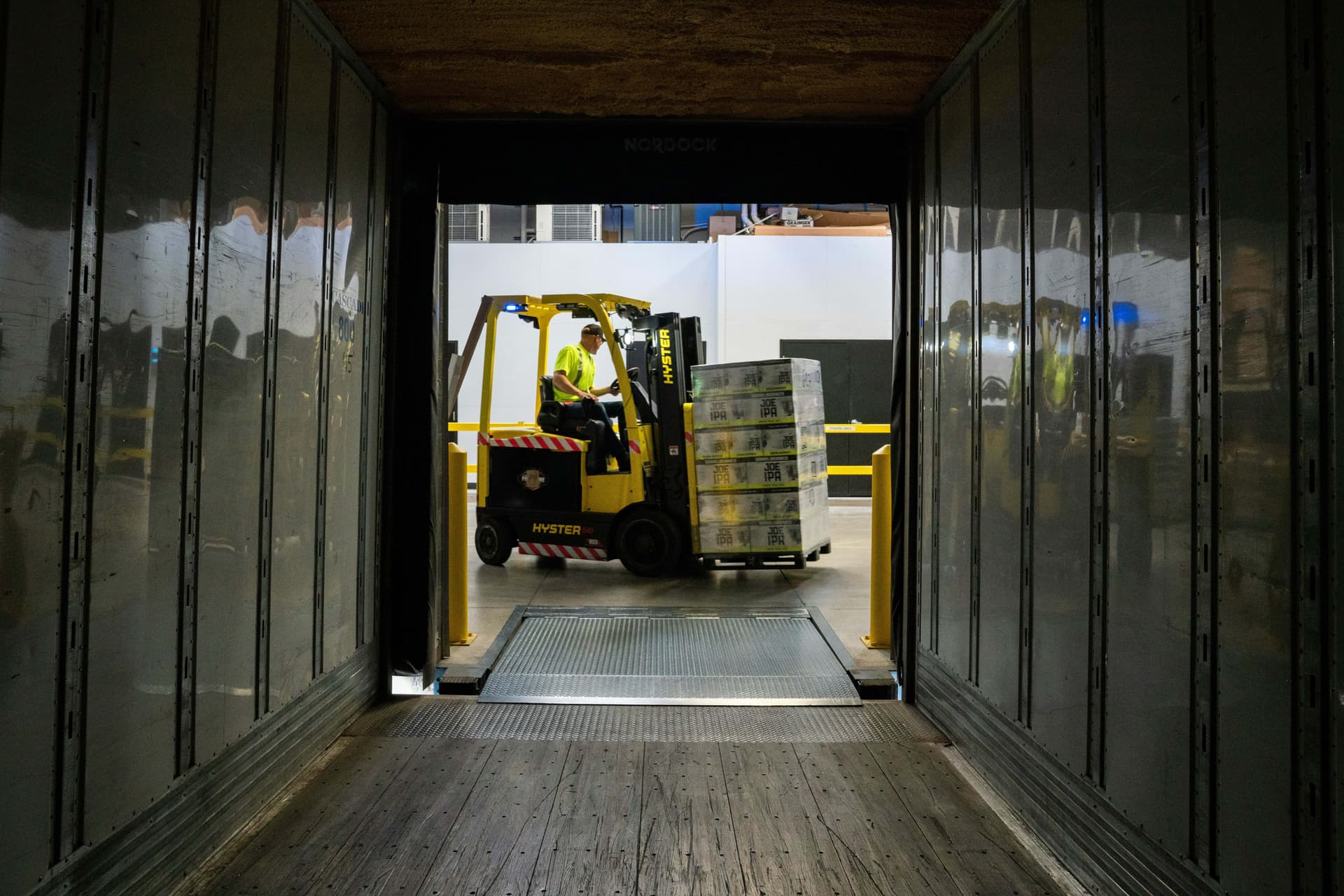 Viewed from inside an empty lorry trailer, a worker drives a yellow forklift carrying a pallet of boxed beer across a loading dock ramp.